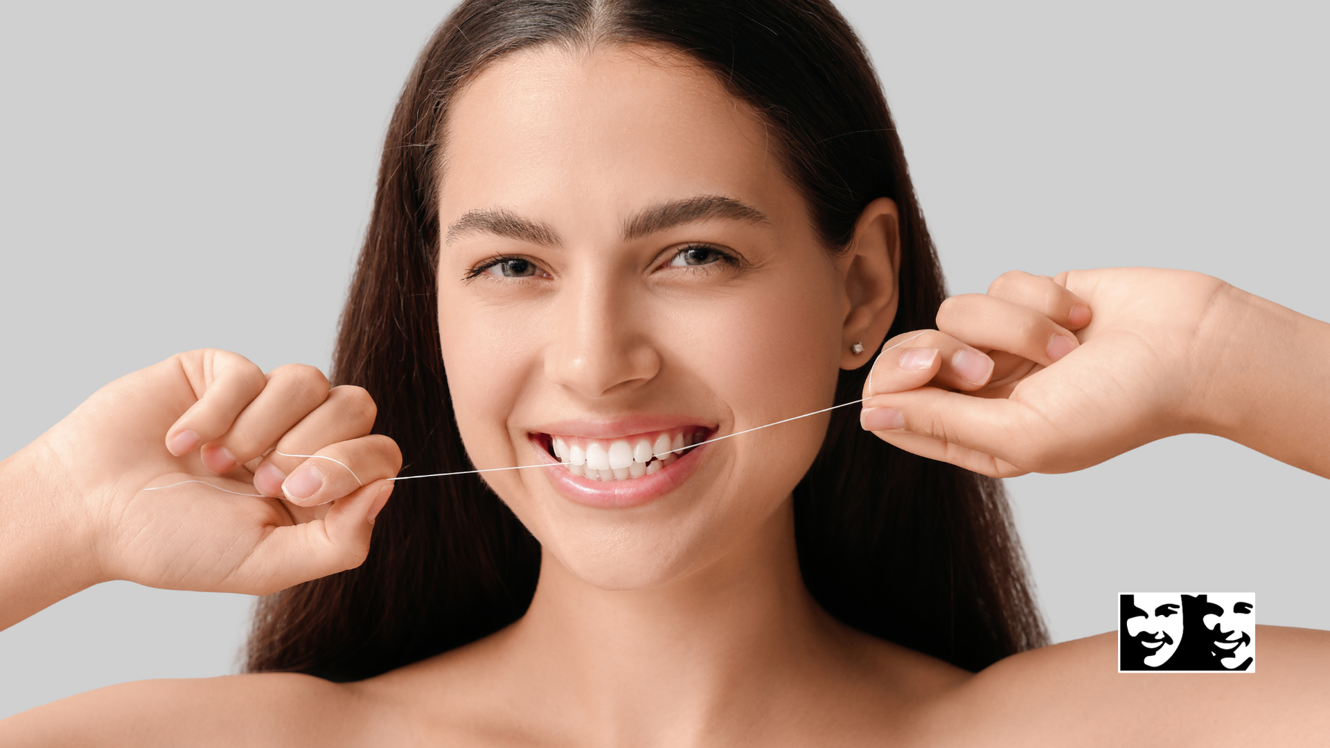 Woman flossing her teeth, smiling widely. Light skin, long dark hair, against a gray background.