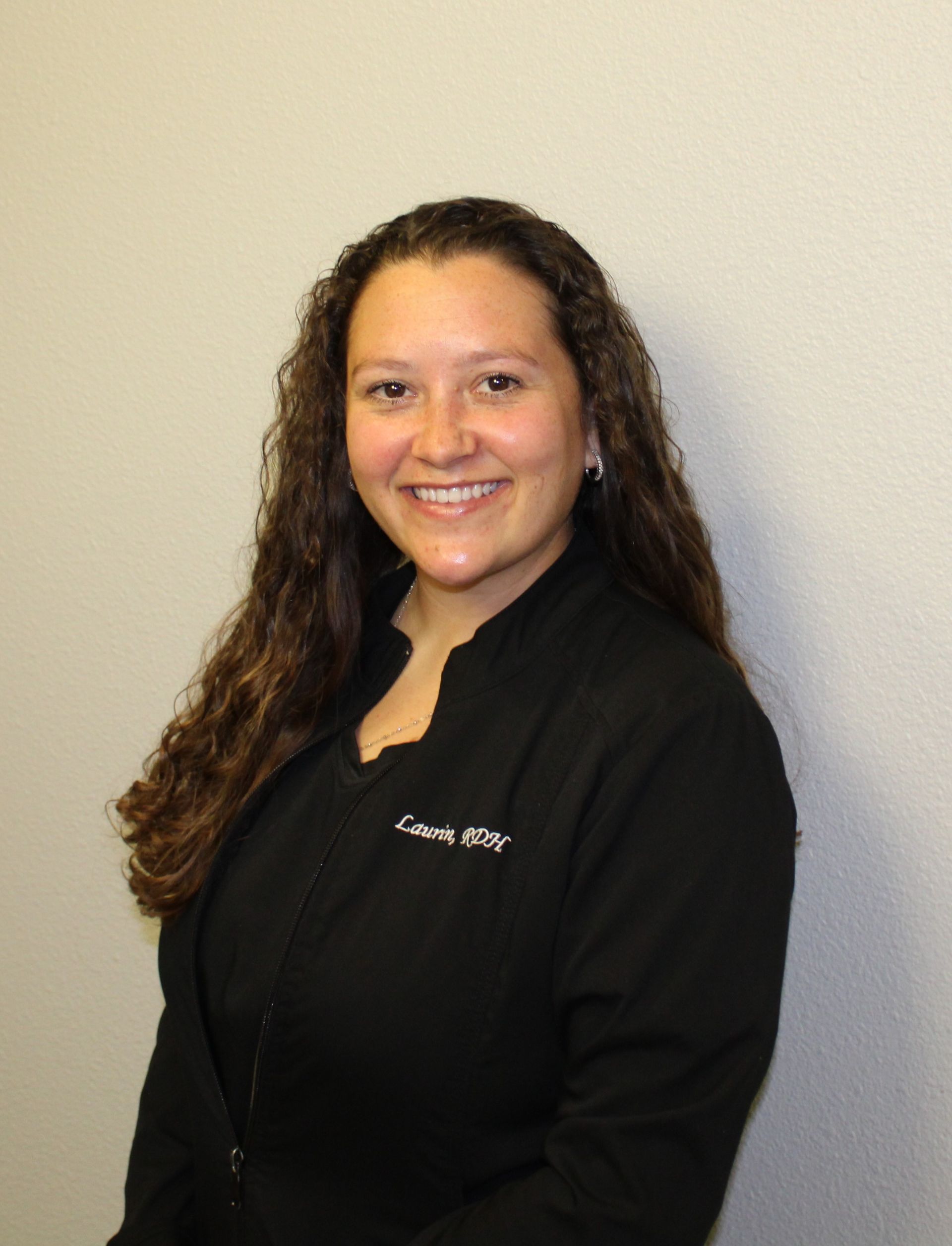Woman with long curly hair, smiling, in black uniform, standing against a white wall.
