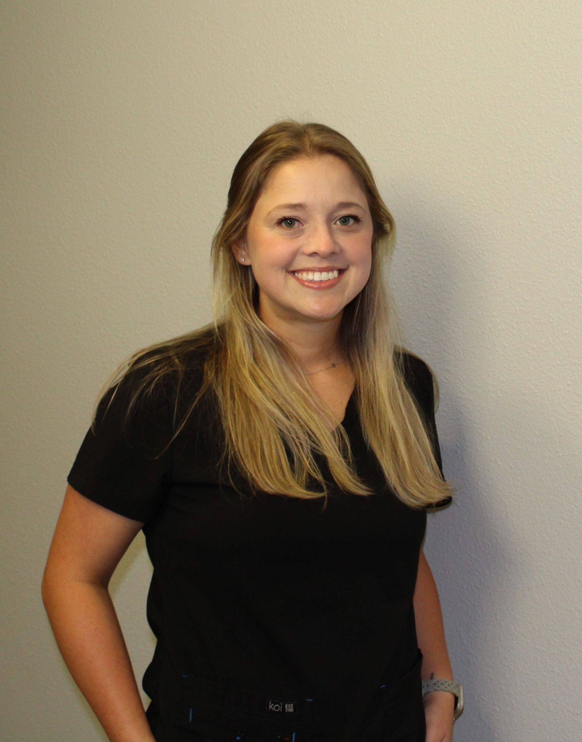 Woman with blonde hair, wearing a black shirt, smiling, standing in front of a white wall.
