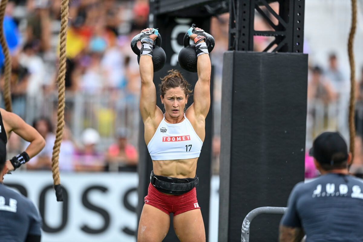 Woman in red shorts and white top, lifting two kettlebells overhead at a CrossFit competition.