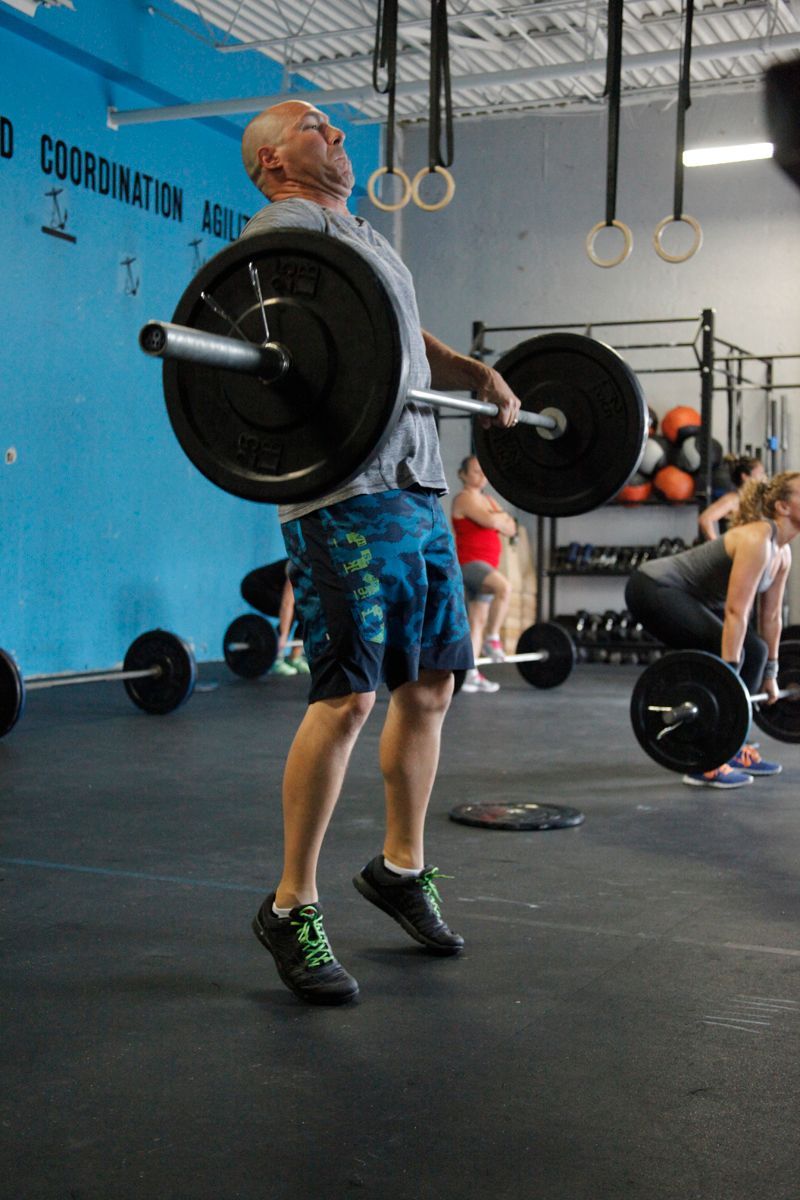 Man lifting barbell in a gym. He wears shorts and a t-shirt, jumping upward. Others exercise in the background.