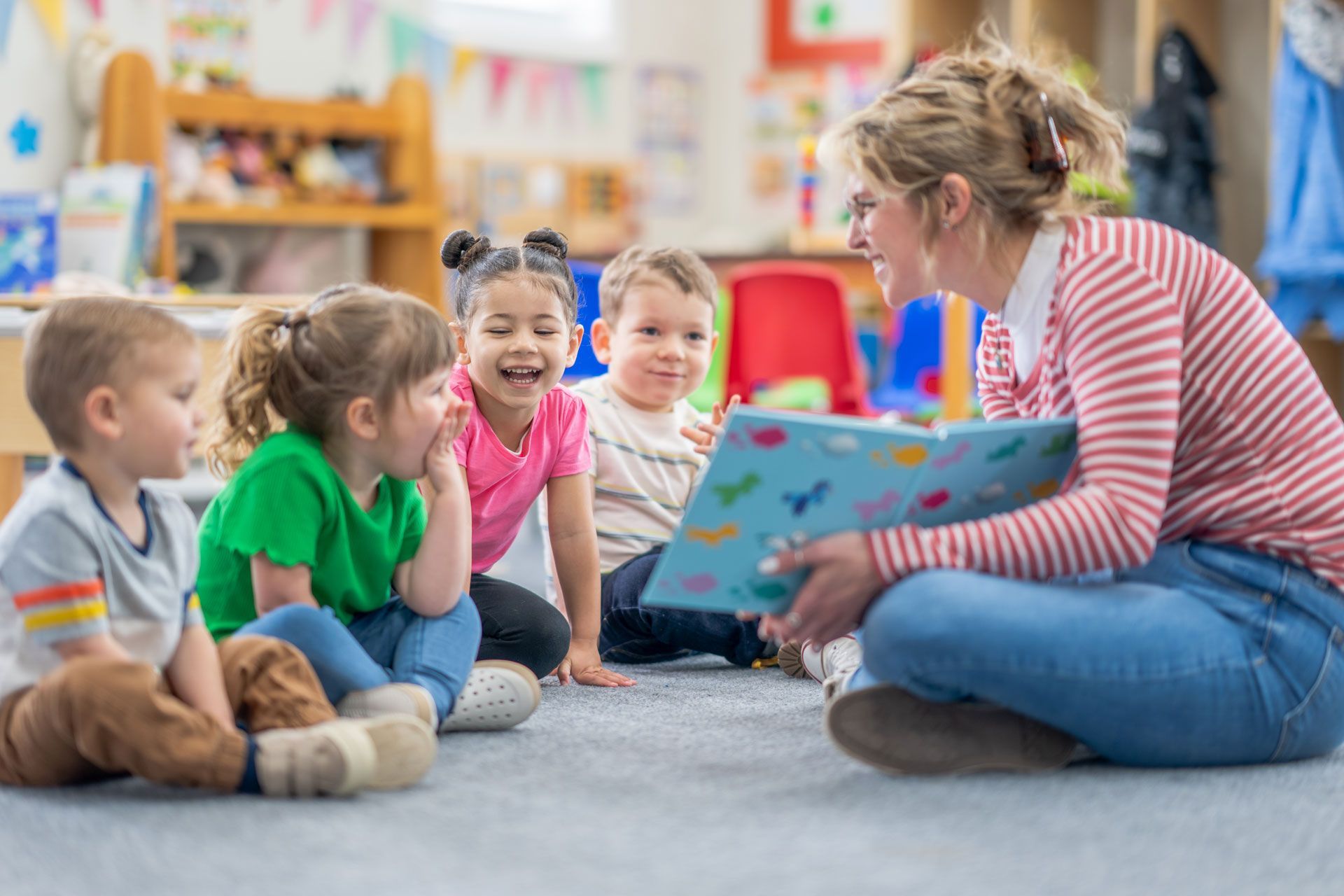 A preschool teacher sits on the floor with a small group of students as she reads them a book.