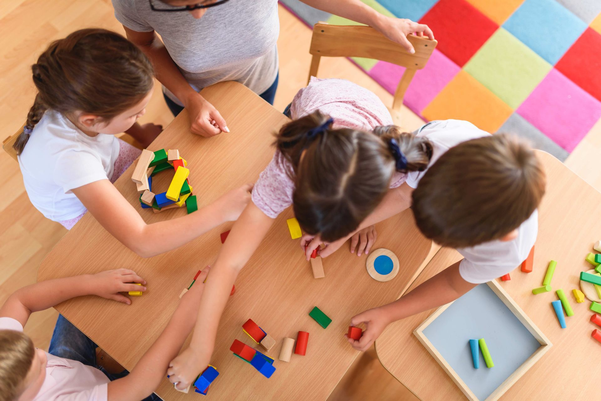 Children playing with colorful wooden blocks at a table while a teacher supervises