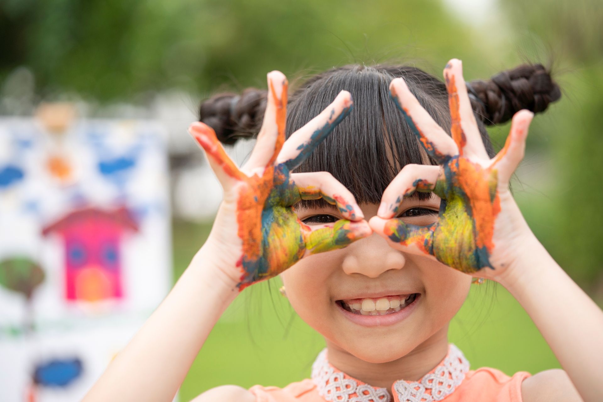 A smiling child with painted hands poses playfully during a colorful outdoor art activity.