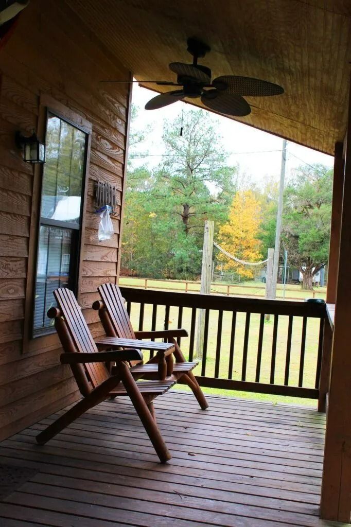 Covered wooden porch with two chairs, ceiling fan, and a view of trees beyond the railing.