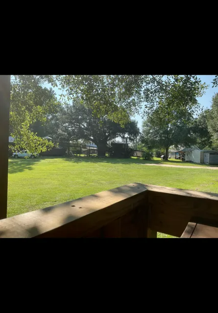 Sunny backyard with green lawn, large trees, and a wooden deck railing in the foreground