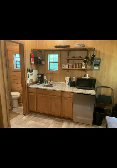 Small rustic kitchen with wooden cabinets, sink, shelves, microwave, and stainless steel dishwasher.