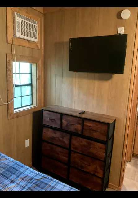 Small wood-paneled bedroom with a wall-mounted TV, dresser, window, and air conditioner.