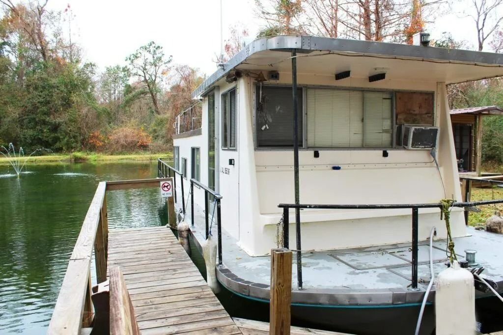 White houseboat docked beside a wooden pier on a calm lake, with trees in the background.