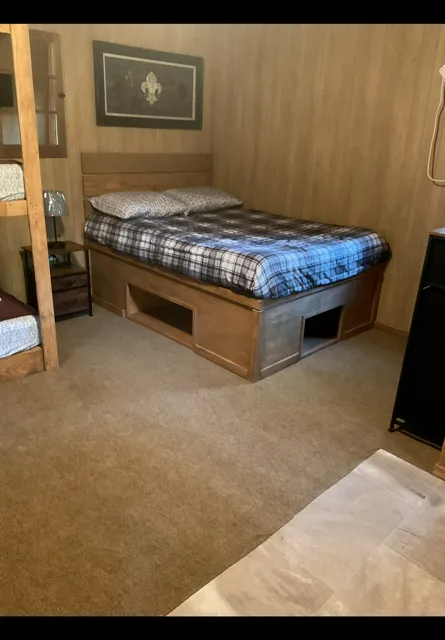 Cozy bedroom with a plaid bed on a wooden platform, paneled walls, carpet, and bunk beds at left.