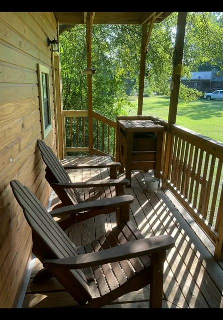 Wooden porch with two Adirondack chairs and a small cabinet overlooking a sunny green yard.