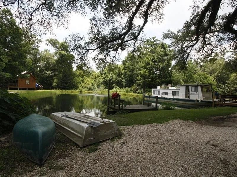 Gravel area beside a pond with boats, a dock, and trees; small buildings in the background.