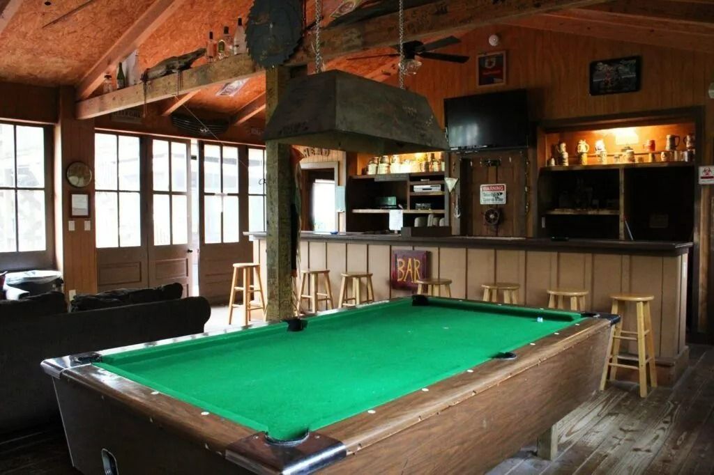 Pool table in a rustic bar with wooden beams, stools, and a large metal hood overhead