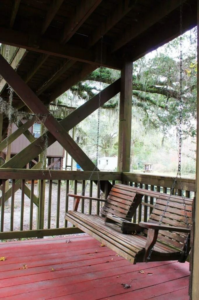 Wooden porch with a red floor, bench swing, and railings overlooking trees in a wooded setting