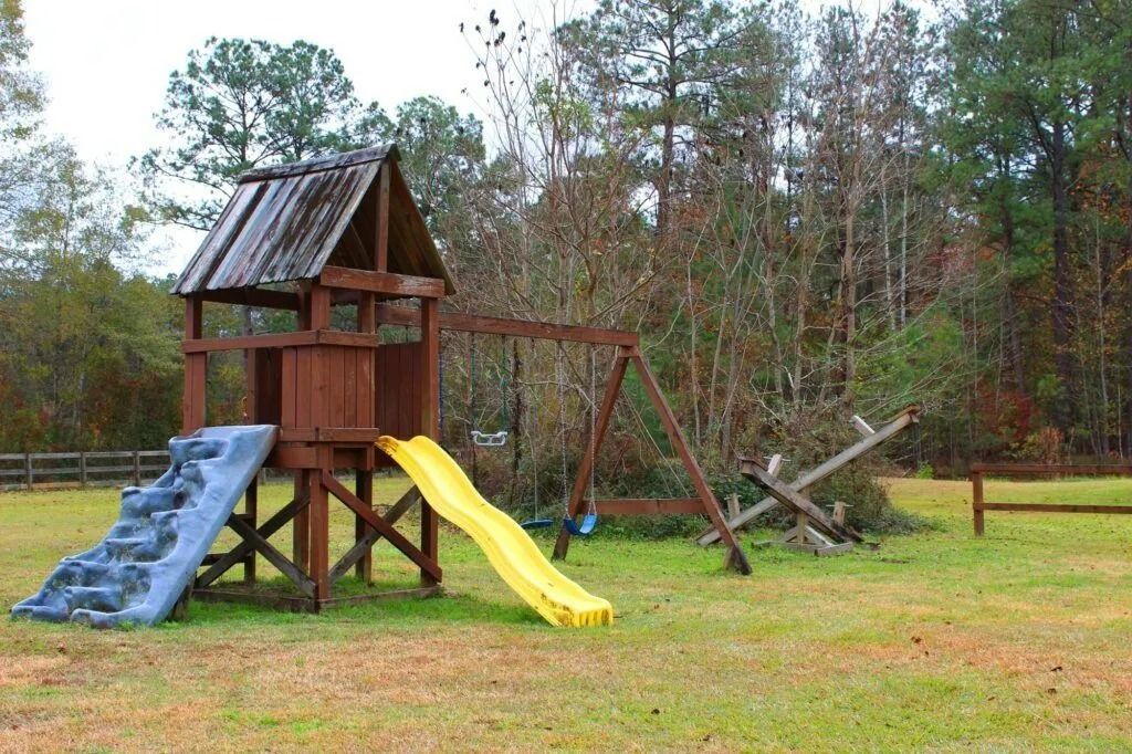 A wooden playground set with blue and yellow slides in a grassy yard near trees.