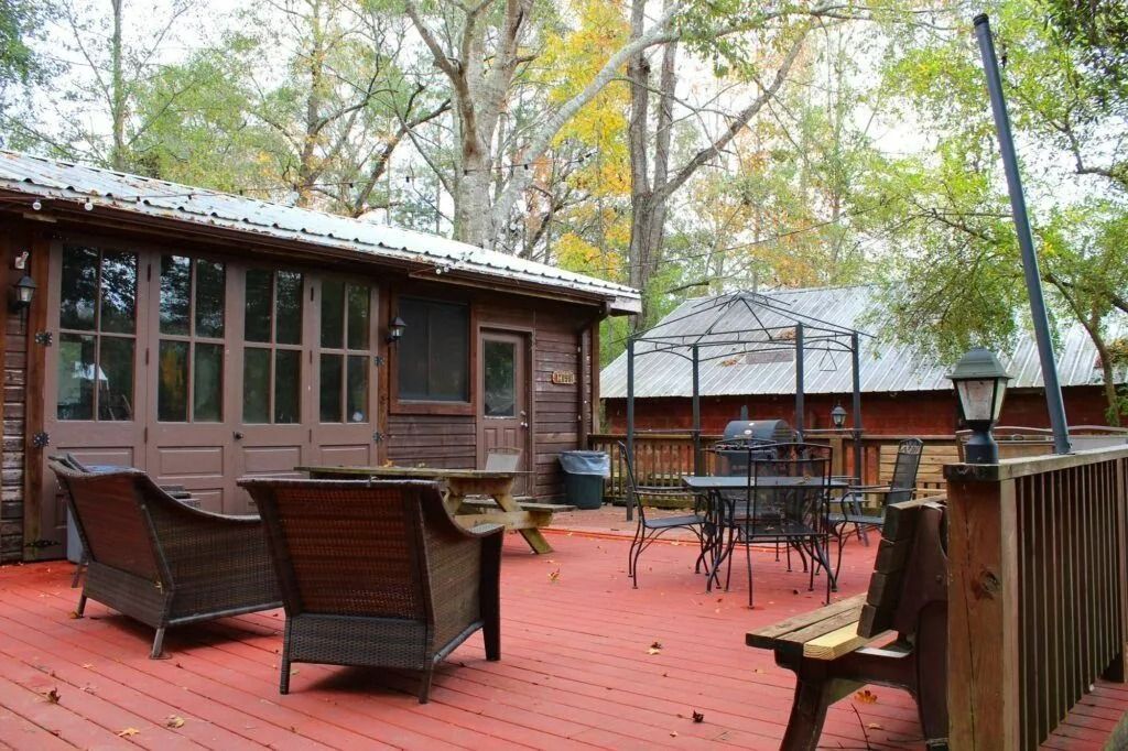 Red wooden patio with wicker chairs and tables beside cabin-like buildings under autumn trees