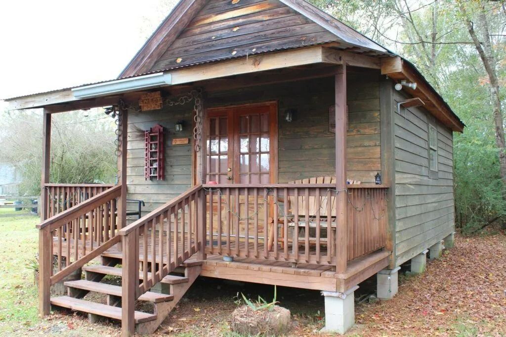 Small rustic wooden cabin with porch, steps, and shingled roof in a wooded setting