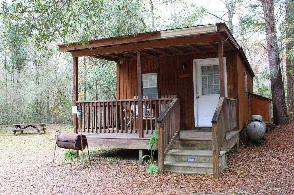 Small rustic cabin with a porch in a wooded area, with a grill and picnic table nearby.