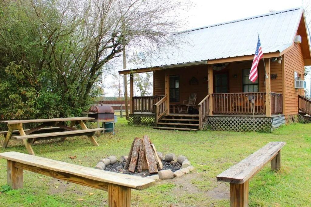 Rustic cabin with a porch, picnic tables, and a fire pit in a grassy yard, with an American flag hanging outside.