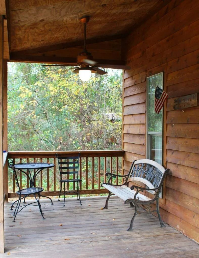 Covered wooden porch with bench, round table, hanging light, and trees beyond the railing.