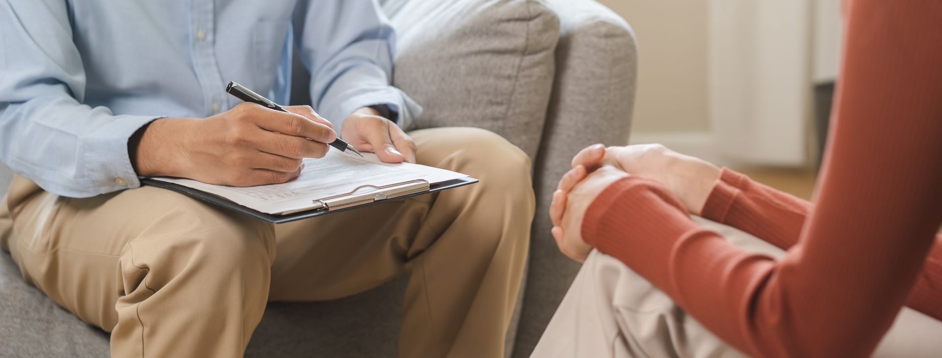 A person in a blue shirt taking notes while another person in a red shirt sits, hands clasped.