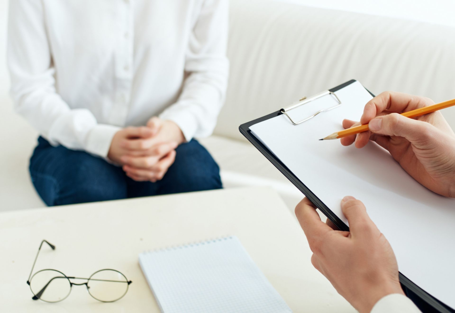 Person in white shirt and jeans sits opposite another person holding a clipboard and writing. Glasses and notepad on table.