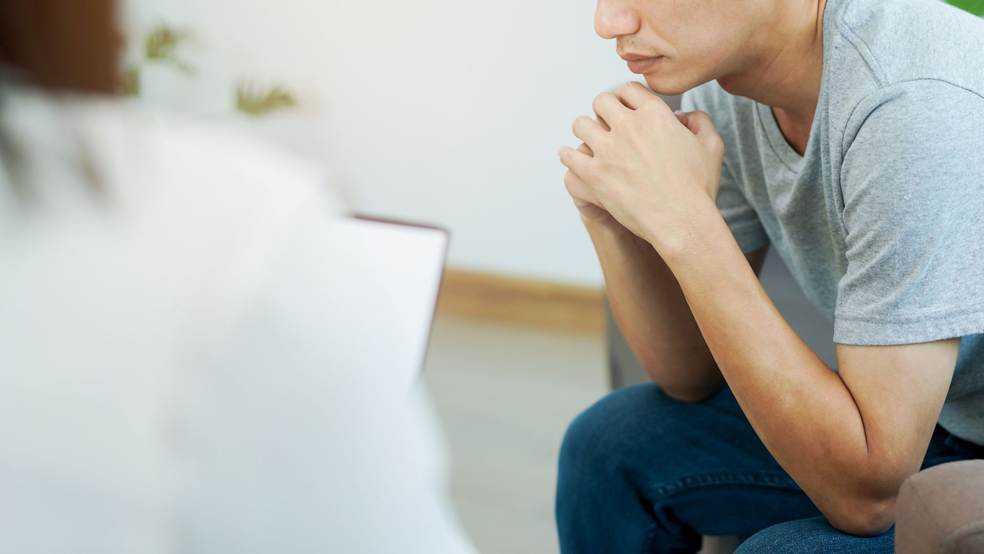 Man in gray shirt sitting, hands clasped, facing person out of view, in a therapy session.