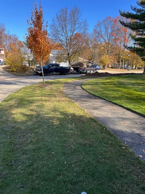 Curved sidewalk through a grassy area, with trees and a truck parked in the background on a sunny day.