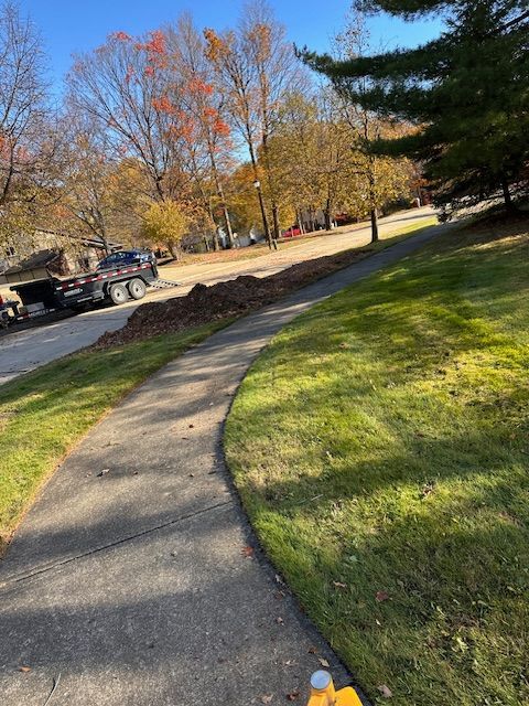 Sidewalk curves along a grassy hill, with street and trees in the background. Leaves and a truck are also present.