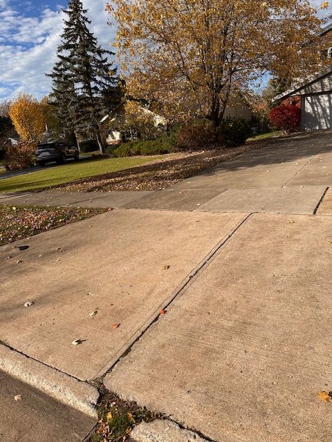 Concrete driveway and sidewalk with fallen leaves, houses, and trees in a residential neighborhood.