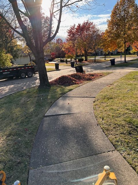 A curved sidewalk in a neighborhood, with fallen leaves and trash cans under autumn trees.