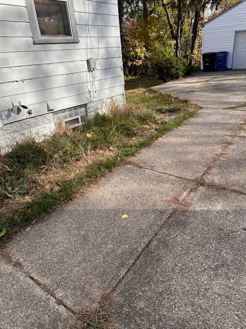 Concrete sidewalk next to a house with overgrown grass, under window.
