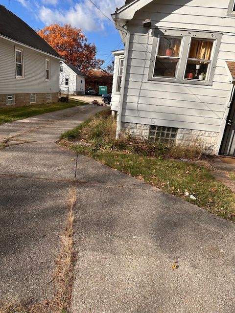 Driveway between two houses, overgrown grass, cloudy sky, fall foliage in the distance.