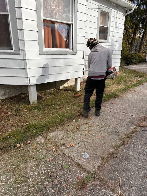 Person using a weed wacker near a house. They are standing on a sidewalk, grass in the foreground.