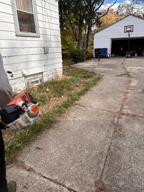 Person using a chainsaw to trim weeds along a concrete driveway and house.