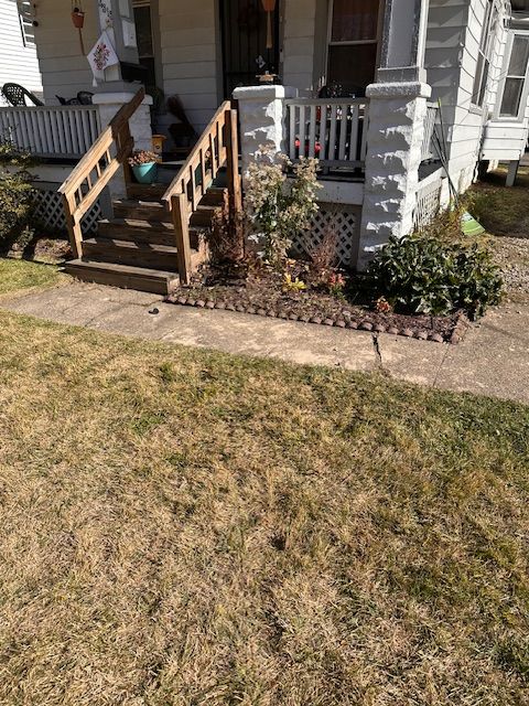 Exterior of a house with a porch and steps. A walkway leads to the house, and there is a small garden bed with plants.