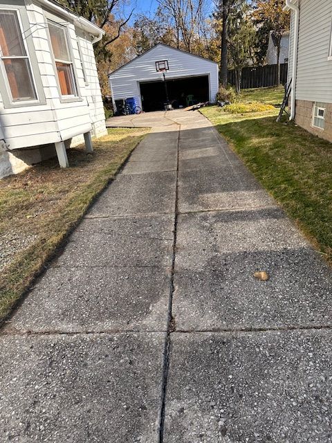 Concrete driveway leading to a garage with a basketball hoop. Buildings and grass on either side.