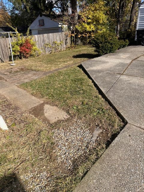 Yard with patches of grass, gravel, and concrete sidewalk. A wooden fence and small garage are in the background.