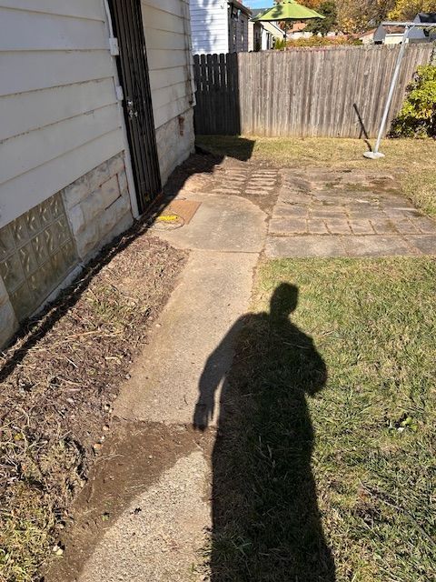 A person's shadow on a concrete walkway leading to a backyard with a wooden fence.