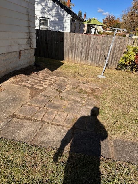 Backyard with a concrete patio, brick pathway, wooden fence, and grass. A shadow falls across the yard.
