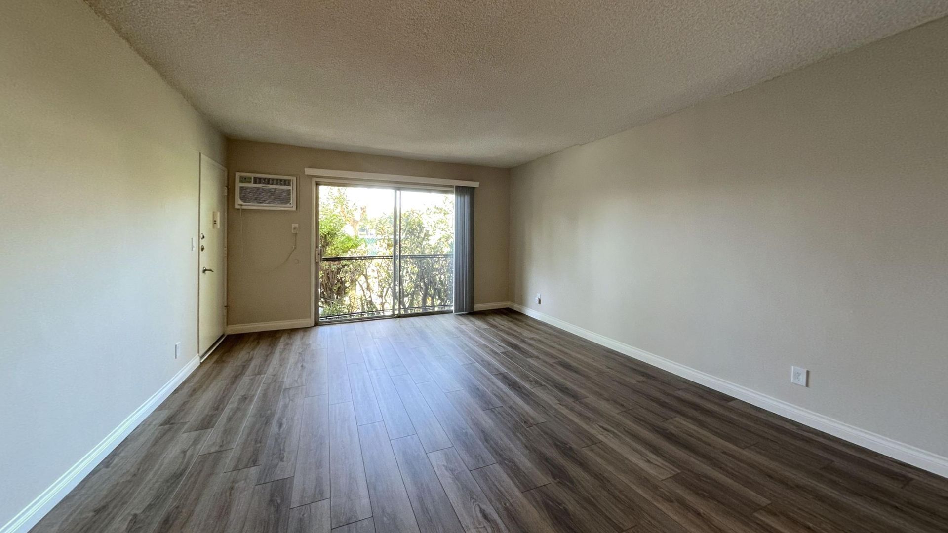 Empty room with wood-look flooring, sliding glass door, and pale walls. An air conditioner is mounted near the door.
