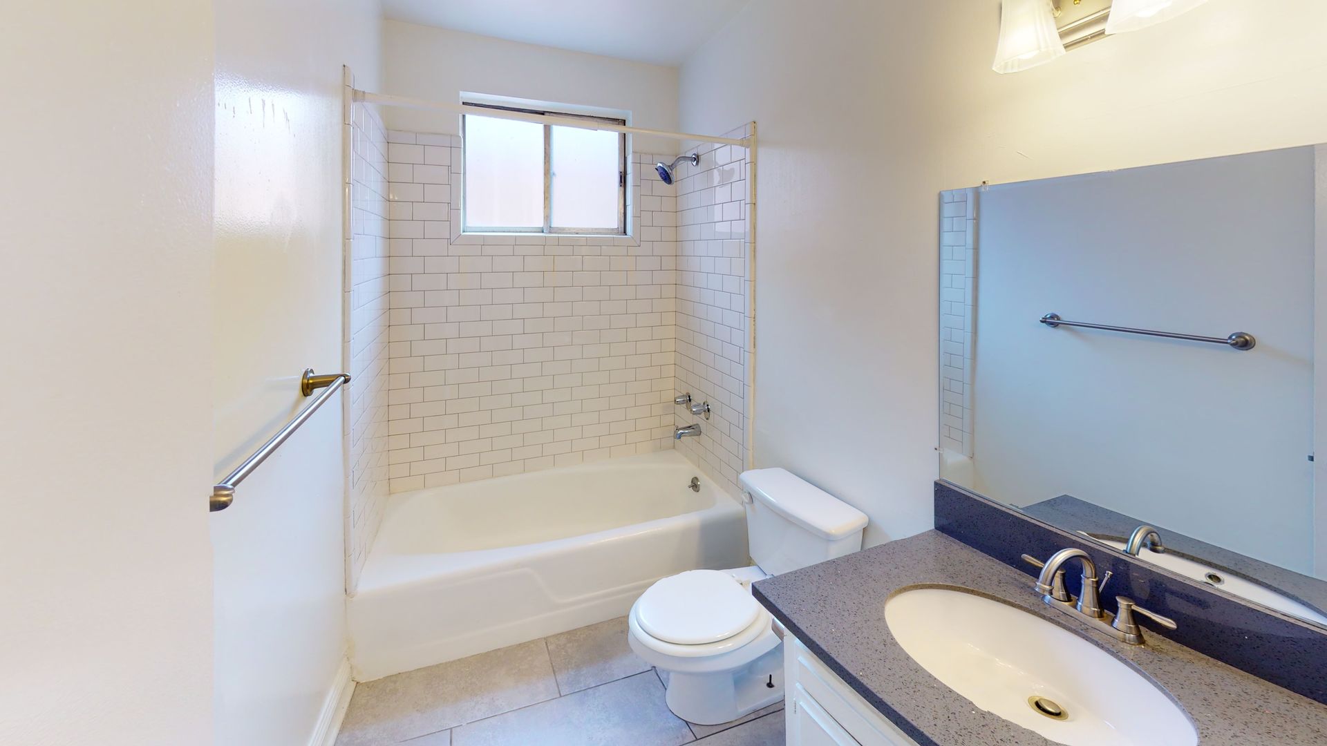 Bathroom with white tub and toilet, gray vanity, showerhead, window, and wall-mounted towel rack.