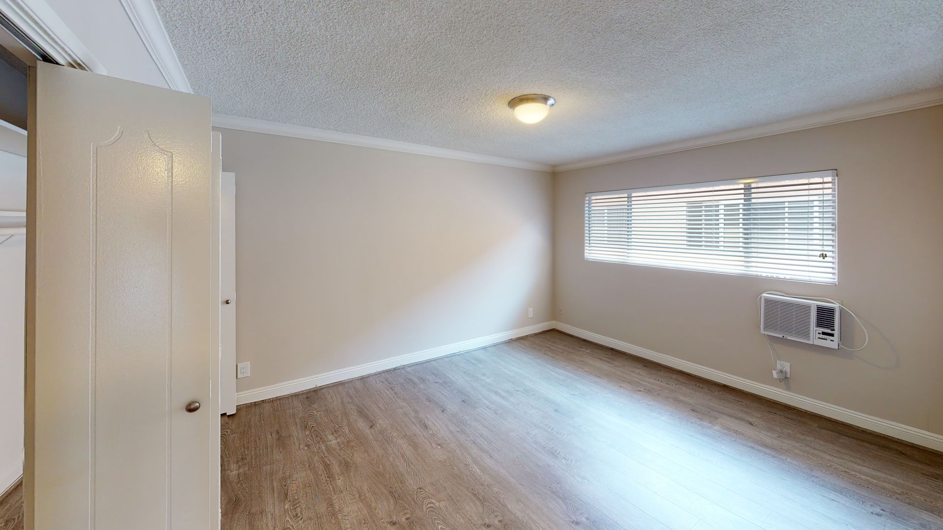 Empty room with wood-look floor, window with blinds, small air conditioner, and white door.