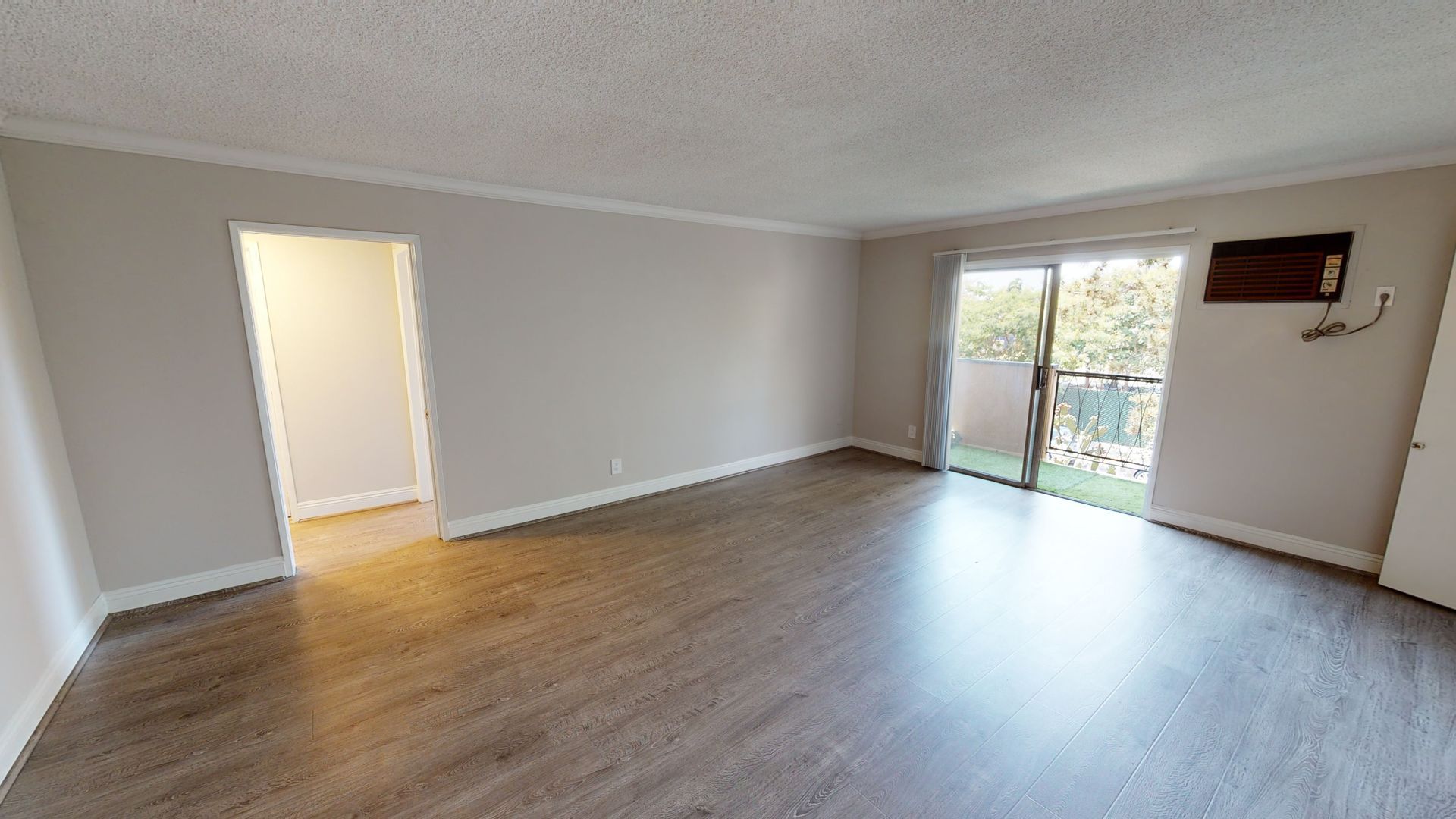 Empty living room with wood-look flooring, door, and sliding door to balcony; neutral walls.
