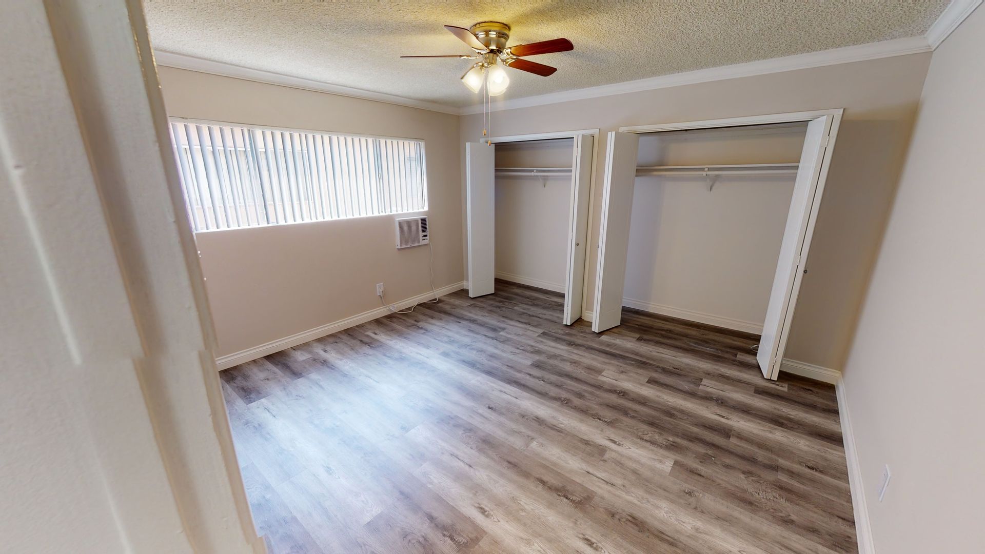 Empty bedroom with wooden floor, two closets, window, and ceiling fan.