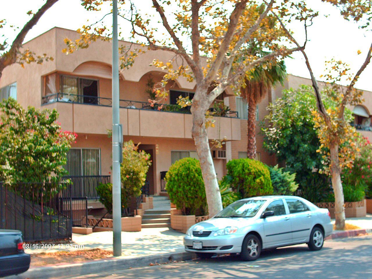 Apartment building with light brown facade and street-parked silver car. Lush greenery and trees line the sidewalk.