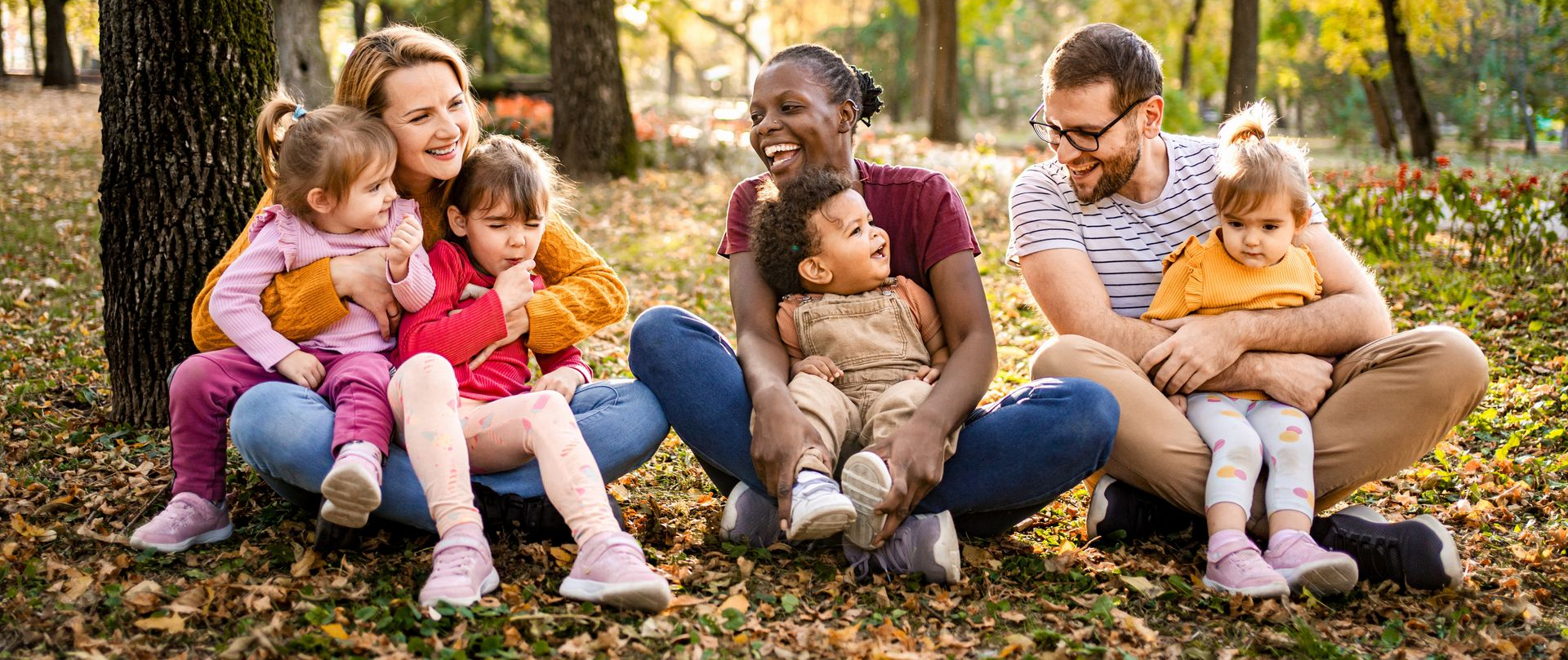 Three families sit in a park, smiling. Each holds a young child. Autumn leaves and trees surround them.