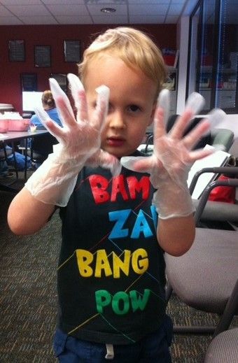 Young child wearing medical gloves, holding hands up. Black shirt with "BAM ZAP BANG POW."