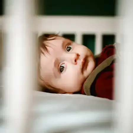 Baby lying in a white crib, looking at the camera with wide eyes.