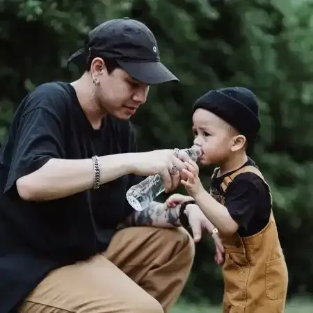 Man kneeling, giving water bottle to a child wearing overalls. Outdoors with green foliage.